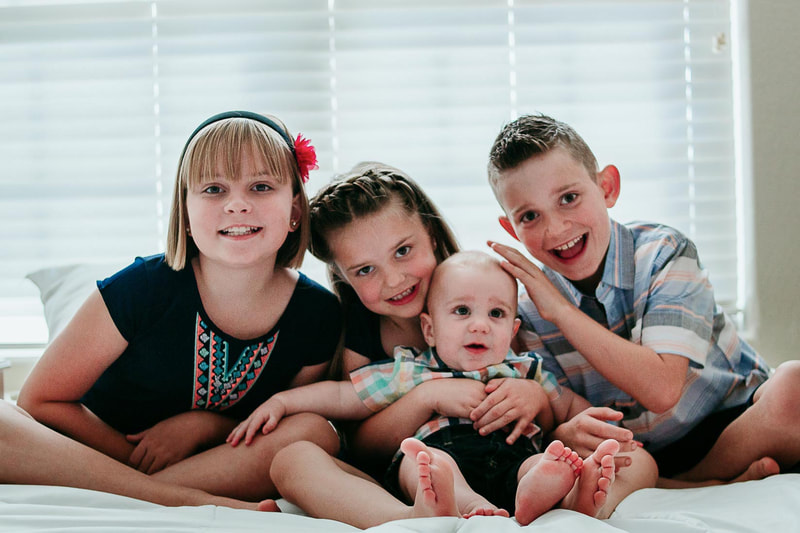 4 children and baby sit on bed and smile at camera.