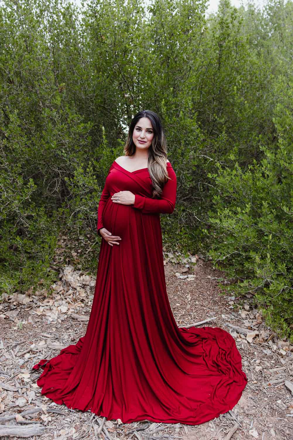 Pregnant woman dressed in red dress in front of green bushes, by your maternity photographer in Albuquerque