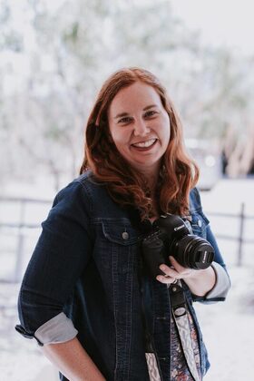 Smiling photo of redhead Anna Cummings wearing a jean jacket and holding a camera.