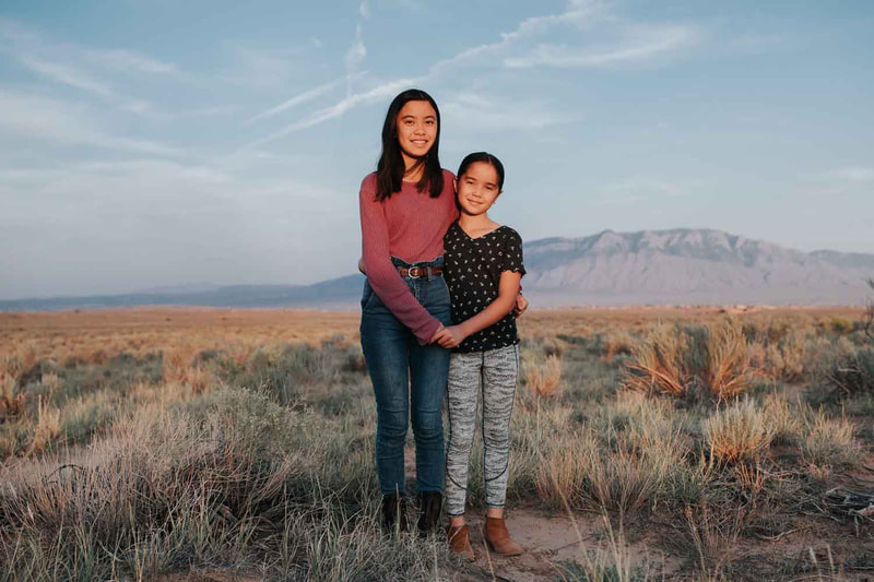 Two young girls hold hands and stand in desert in front of mountains.