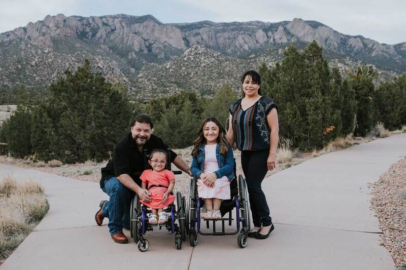 Mom and daughter sit in wheelchairs with grandma and grandpa in front of mountains.