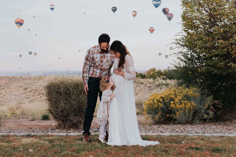 pregnant coupld stand in front of sky with hot air balloons as young daughter kisses mom's pregnant belly.