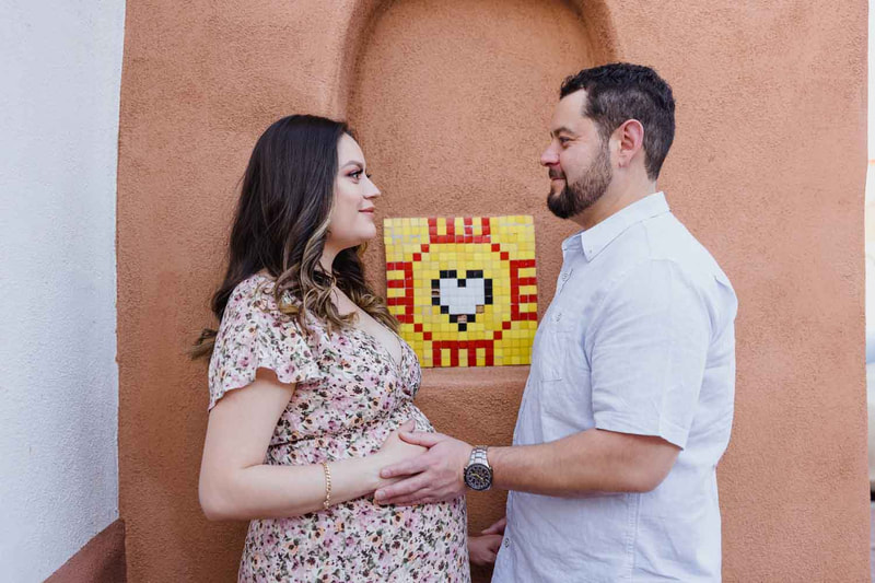 pregnant couple stand in front of zia symbol with heart.