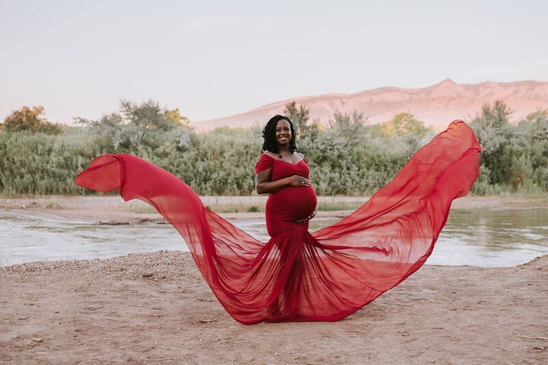 Pregnant mother stands in red dress in front of rvier and mountains as sides of dress fly up