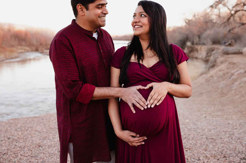 Pregnant couple in red stand together and have hands on belly in heart shape.