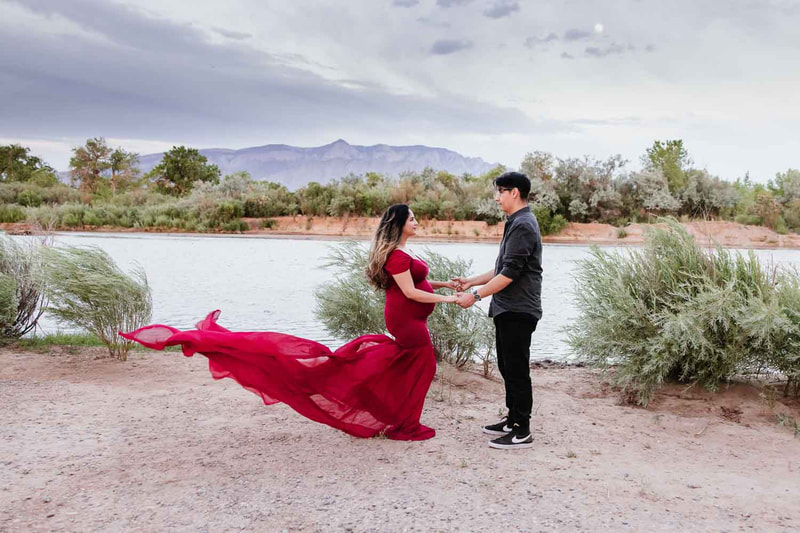 Couple stand and hold hands in front of river as pregnant mom's red dress flies up behind her