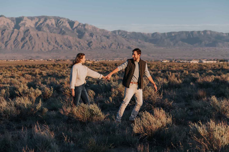 Couple hold hands and walk in front of mountains.