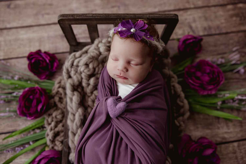 Newborn girl wrapped in purple laying on small bed with purple flowers around.