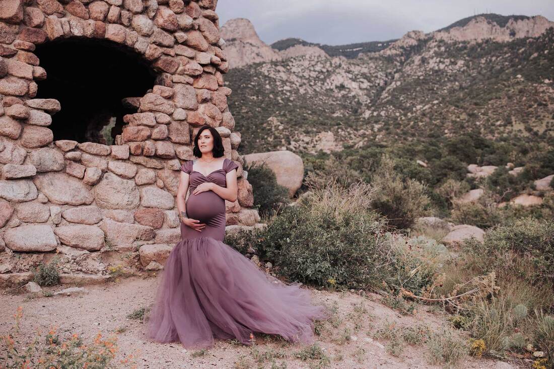 Pregnant mother stands in front of stone cabin wearing flowing purple gown.