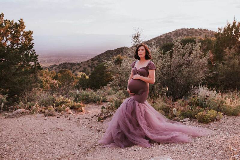Pregnant mother to be stands in front of mountains in a purple gown holding her belly.