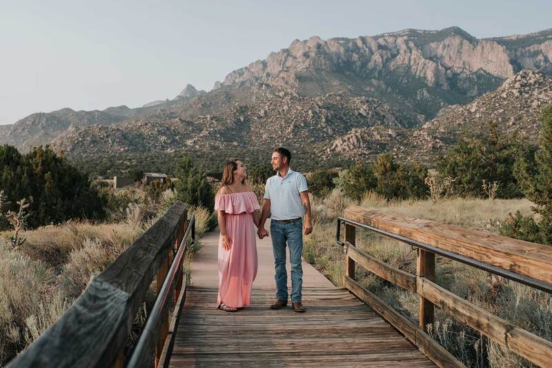 Couple holds hands and walk over bridge in front of mountains.