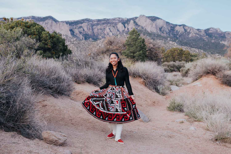 Female senior stands in front of mountains while wearing native american skirt.