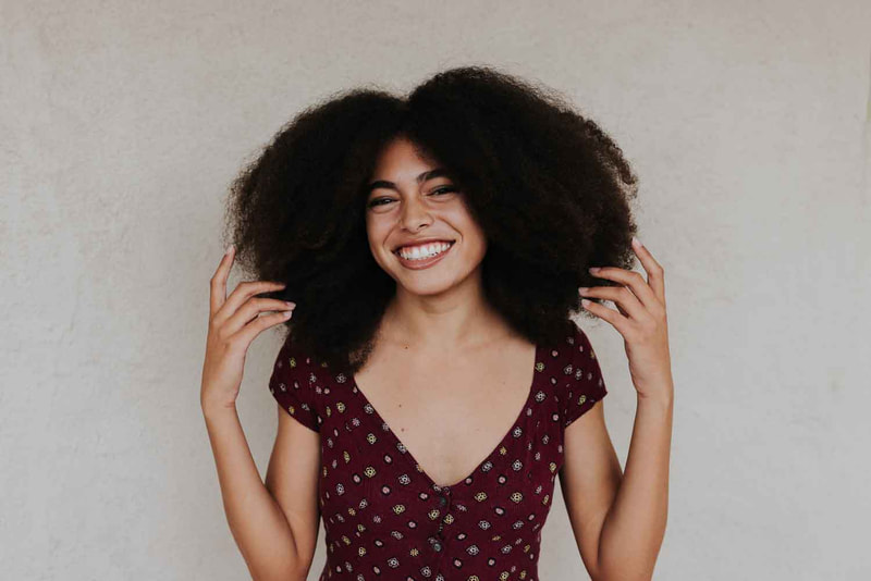 Female senior stands in front of white wall and holds hands on either side of afro.