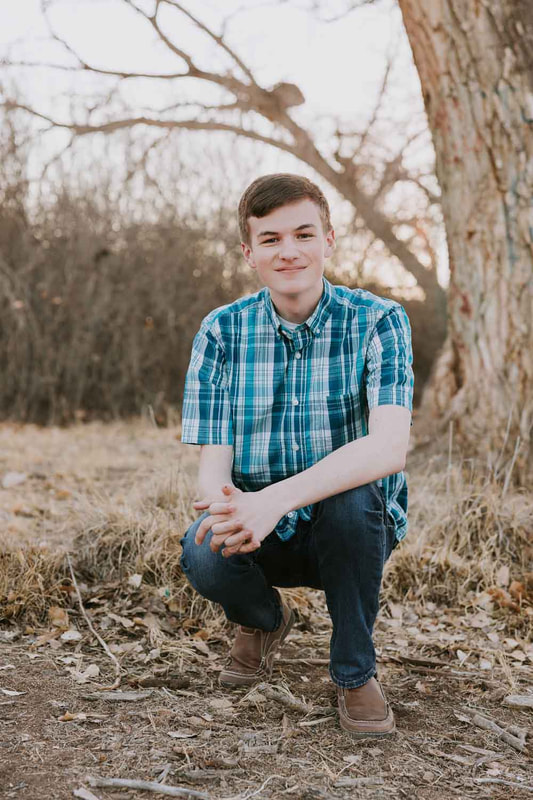 Senior boy smiles at camera while crouching in front of tree.
