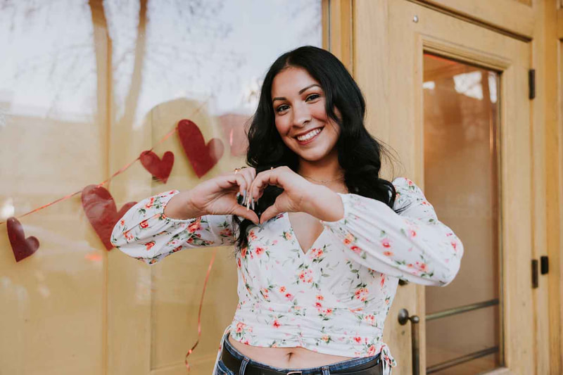 Female senior stands in front of window with hearts and makes a heart symbol with hands.