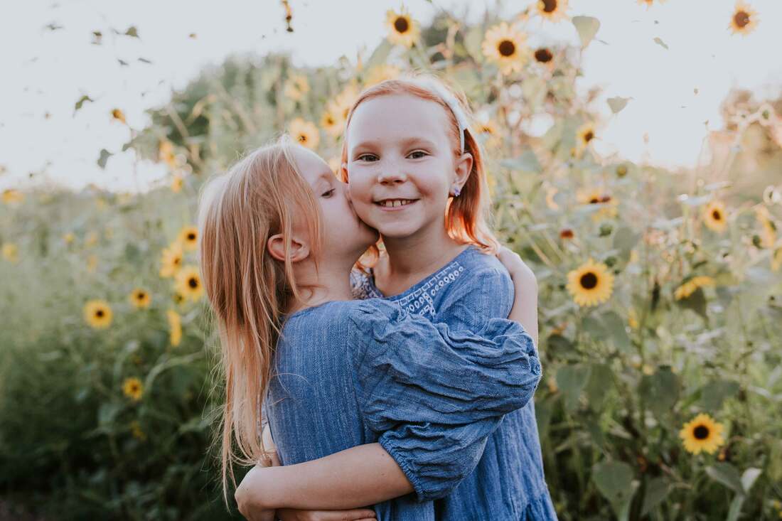 Younger sister kissing older sister on cheek in front of sunflowers
